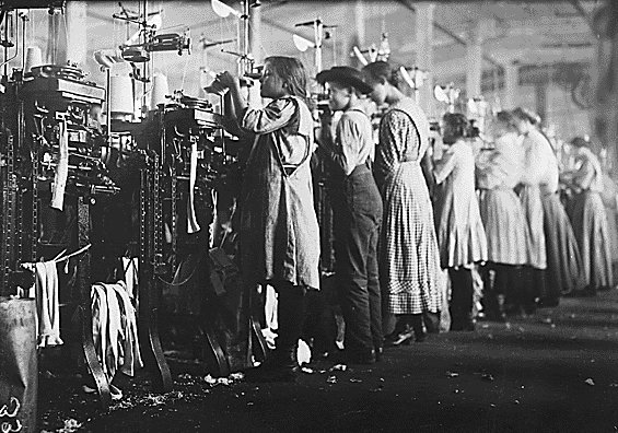 Fotografía blanco y negro de mujeres y niños realizando diversas actividades en una fábrica textil en la época de la Revolución Industrial.
